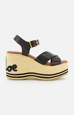 The CHLOÉ Chlóe Sand Wedge Heel is a black leather sandal with crisscross straps, ankle buckle, espadrille-style jute sole with black stitching, wavy heel design, and classic wedge heel flair.