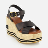 The Chlóe Sand Wedge Heel by CHLOÉ is a black leather sandal with crisscross straps, gold buckle, woven straw heel with black stripes, a small side bow, and signature logo detailing.