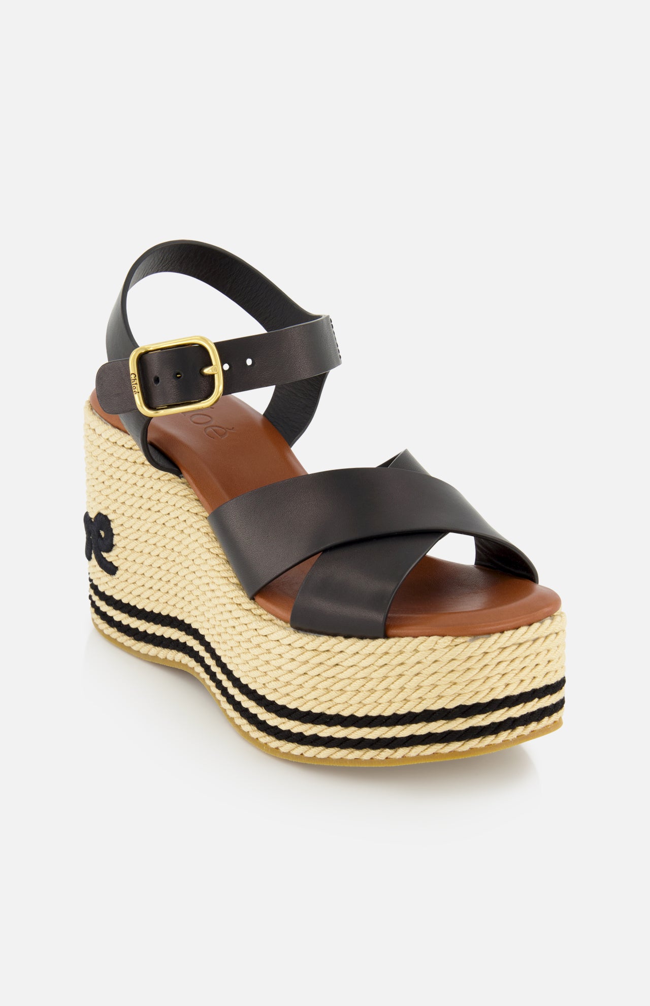 The Chlóe Sand Wedge Heel by CHLOÉ is a black leather sandal with crisscross straps, gold buckle, woven straw heel with black stripes, a small side bow, and signature logo detailing.
