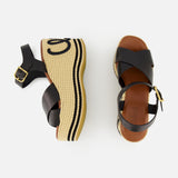The CHLOÉ Chlóe Sand Wedge Heel features black leather straps, ankle buckles, and woven beige platforms with black wavy trim and stitched details. One sandal is shown from the side, the other from above.