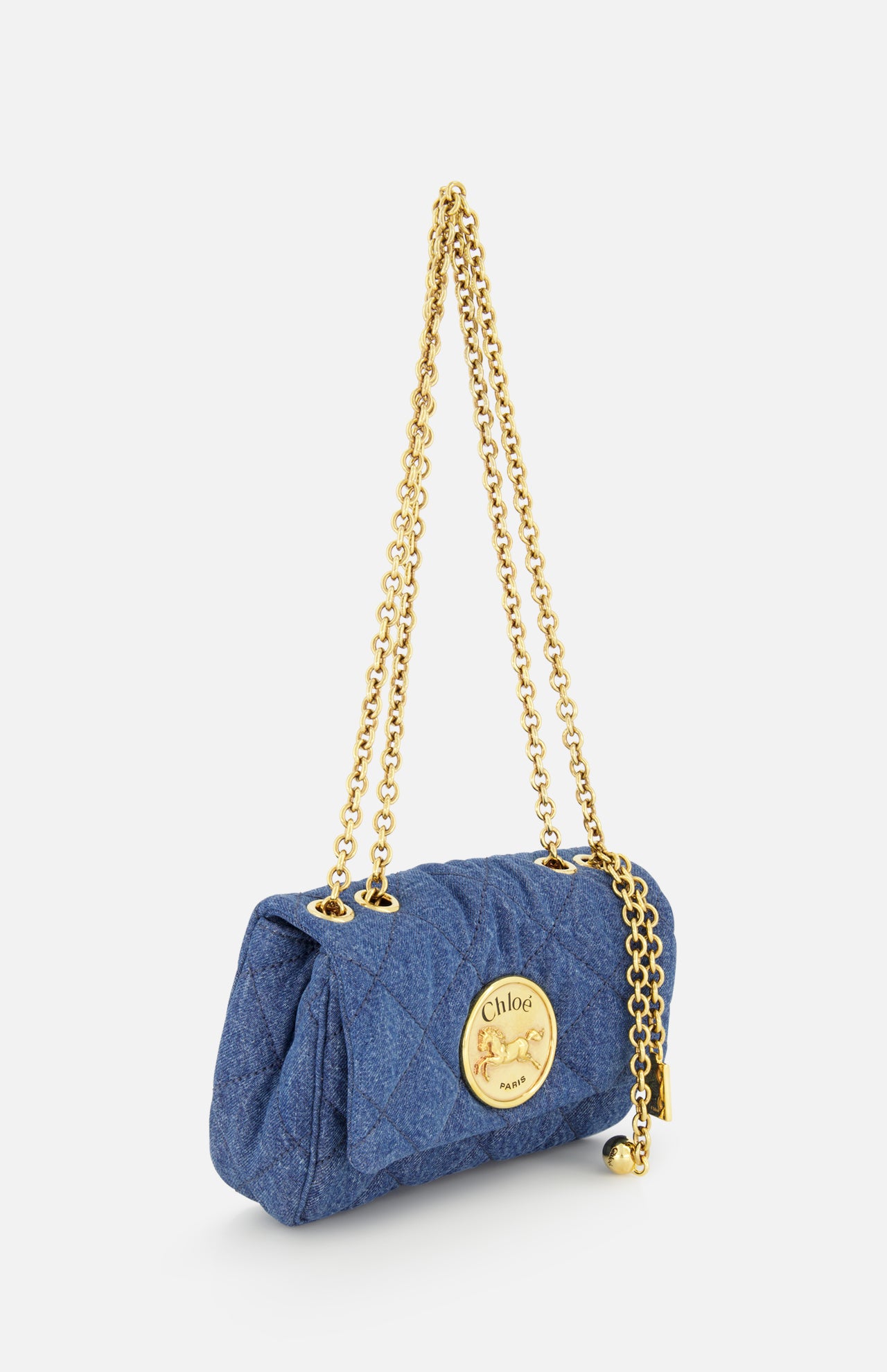 The CHLOÉ Quilted Denim Heritage Bag features a blue quilted denim design, a gold chain strap, and a prominent gold circular Chloé Paris logo on the front, shown against a white background.