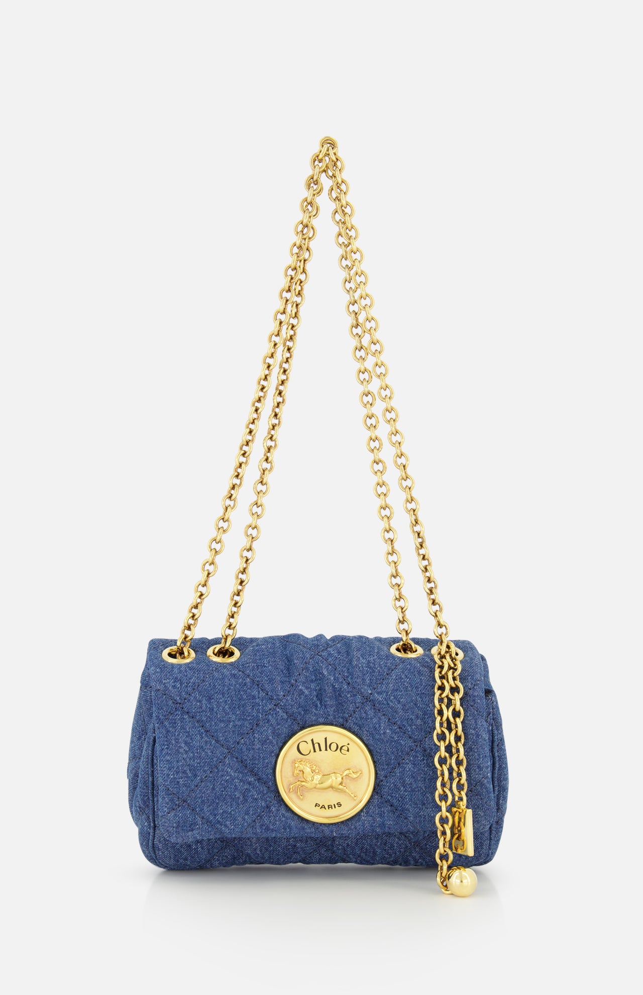 The CHLOÉ Quilted Denim Heritage Bag is a small blue quilted denim shoulder bag with a gold chain strap and a bold round gold emblem featuring the Chloé logo and horse on the front.