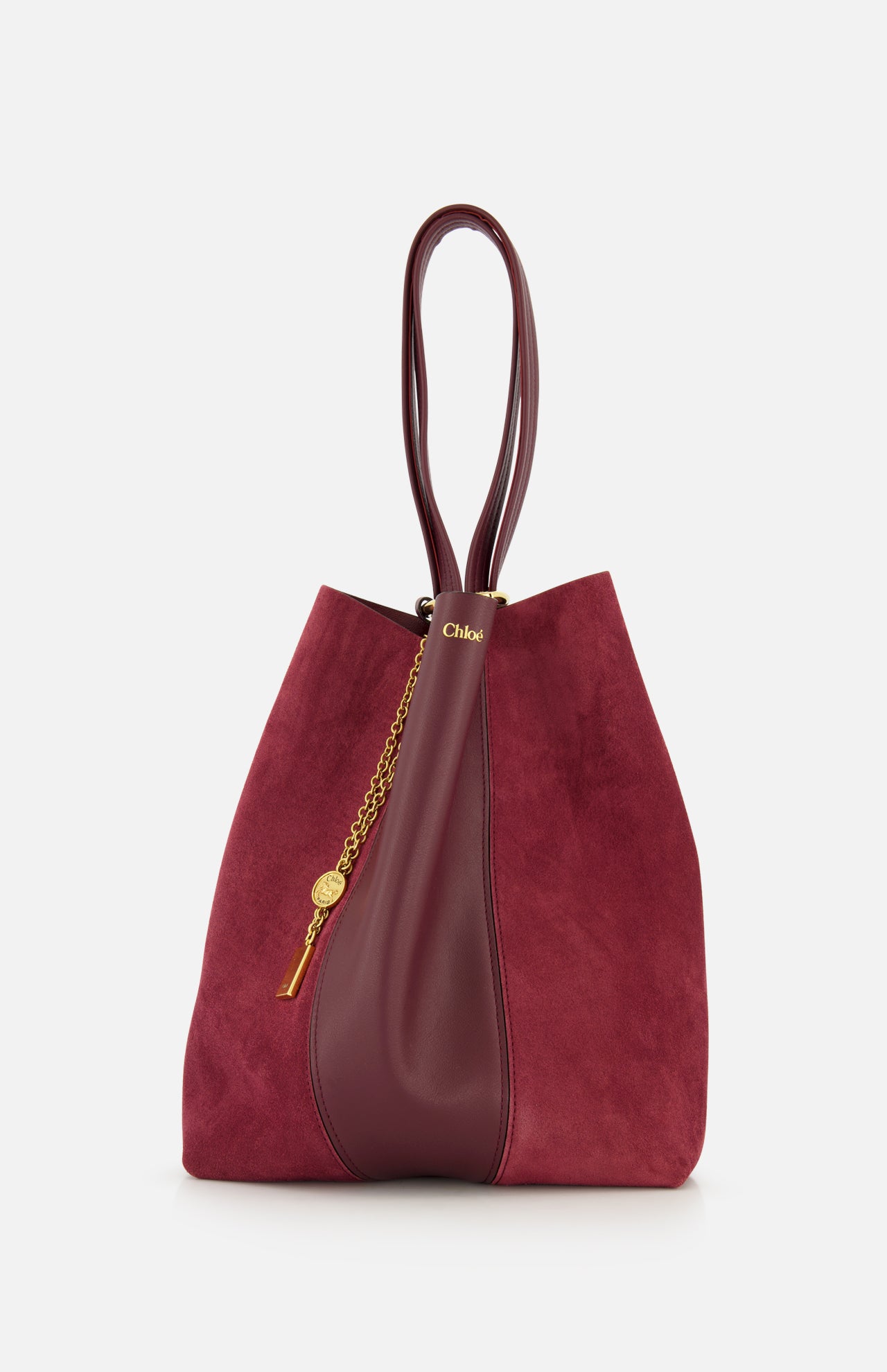 The CHLOÉ Leather and Suede Spin Bag is a burgundy tote featuring double handles, crafted from a mix of suede and leather. It showcases a stylish removable bag chain, an elegant gold medallion detail, and a subtle gold logo near the top.