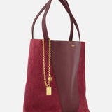 The CHLOÉ Leather and Suede Spin Bag is a sophisticated tote, crafted in rich burgundy leather with suede side panels and a smooth leather center, reflecting the brand’s signature elegance. It features double handles and a removable chain accented with a small gold padlock charm, presented against a plain white background.