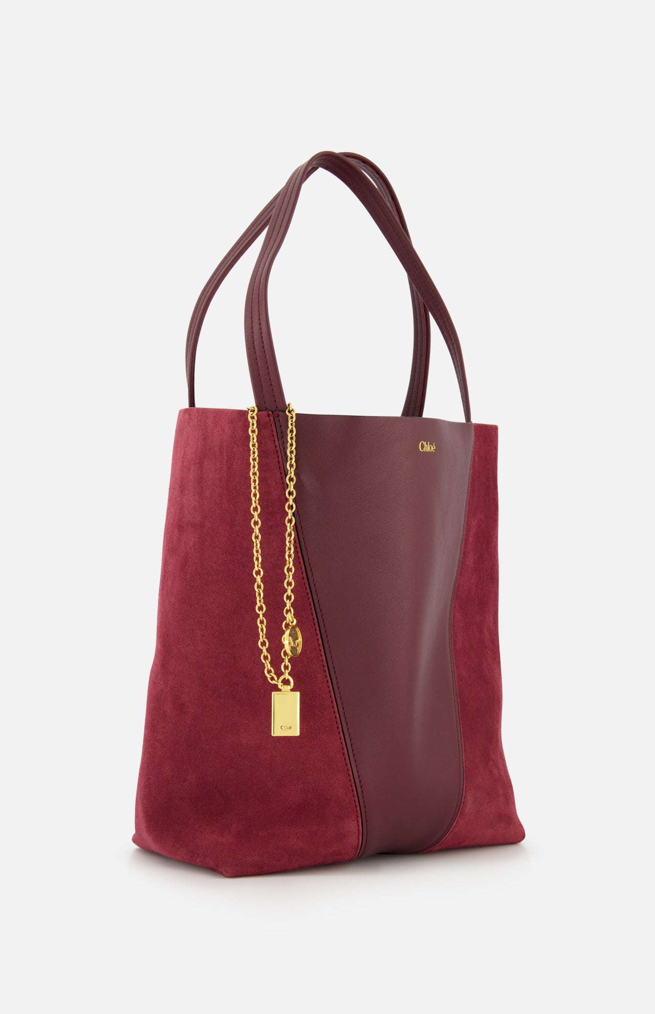 The CHLOÉ Leather and Suede Spin Bag is a sophisticated tote, crafted in rich burgundy leather with suede side panels and a smooth leather center, reflecting the brand’s signature elegance. It features double handles and a removable chain accented with a small gold padlock charm, presented against a plain white background.