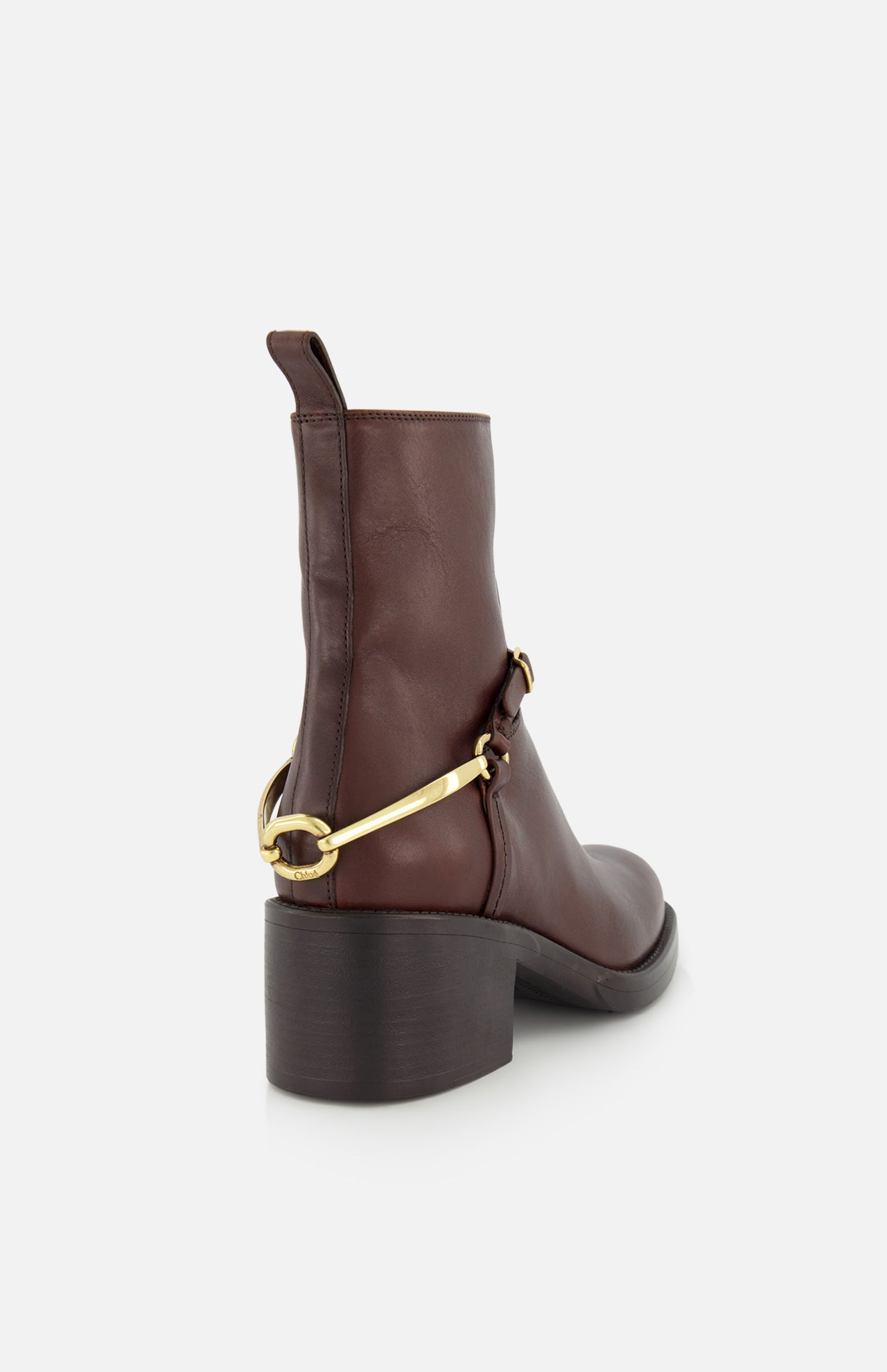 The Dakota Riding Boot by CHLOÉ is a dark brown leather ankle boot with a chunky heel, equestrian-inspired gold hardware at the back, and a pull tab, shown from the rear against a white background.