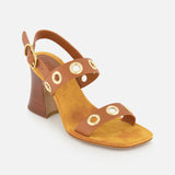The Janis Slingback Grommet Heels by CHLOÉ are ginger brown leather sandals featuring a medium wooden block heel and two straps detailed with gold circular grommets and a gold buckle. These CHLOÉ leather slingbacks have a yellow suede insole and are photographed against a plain white background.