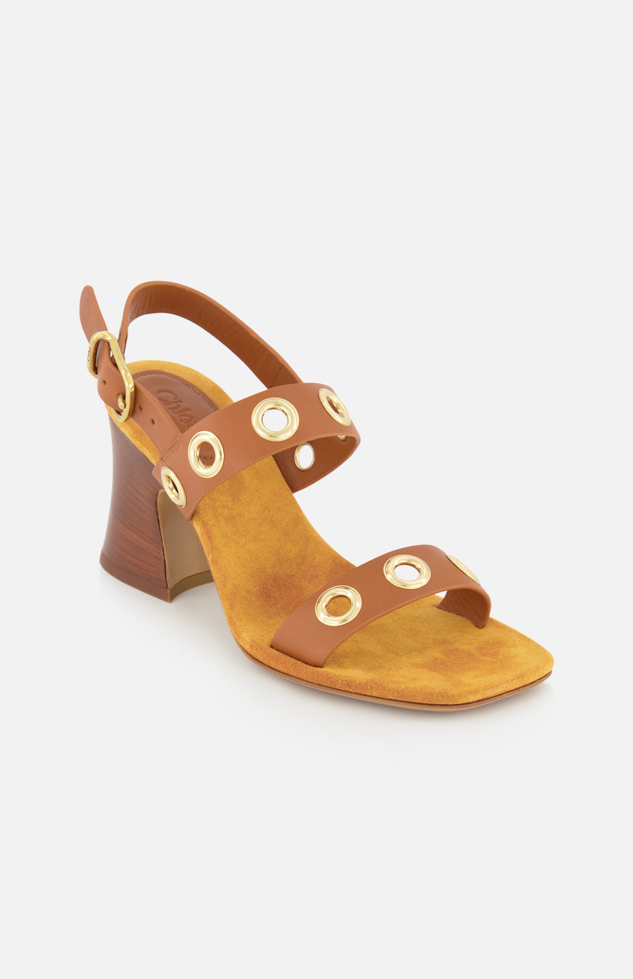 The Janis Slingback Grommet Heels by CHLOÉ are ginger brown leather sandals featuring a medium wooden block heel and two straps detailed with gold circular grommets and a gold buckle. These CHLOÉ leather slingbacks have a yellow suede insole and are photographed against a plain white background.