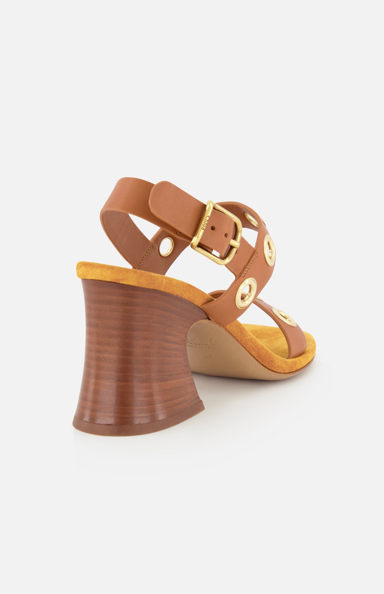 The Janis Slingback Grommet Heels by CHLOÉ—a pair of ginger brown leather slingback sandals with gold buckle and rounded gold grommet accents, featuring a flared wooden block heel and a tan insole—are shown from the back left side against a white background.