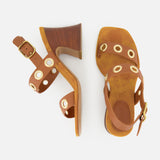 The Janis Slingback Grommet Heels by CHLOÉ are ginger brown sandals, crafted from supple leather with gold grommet accents, ankle straps, and chunky wooden block heels, displayed on a white background.
