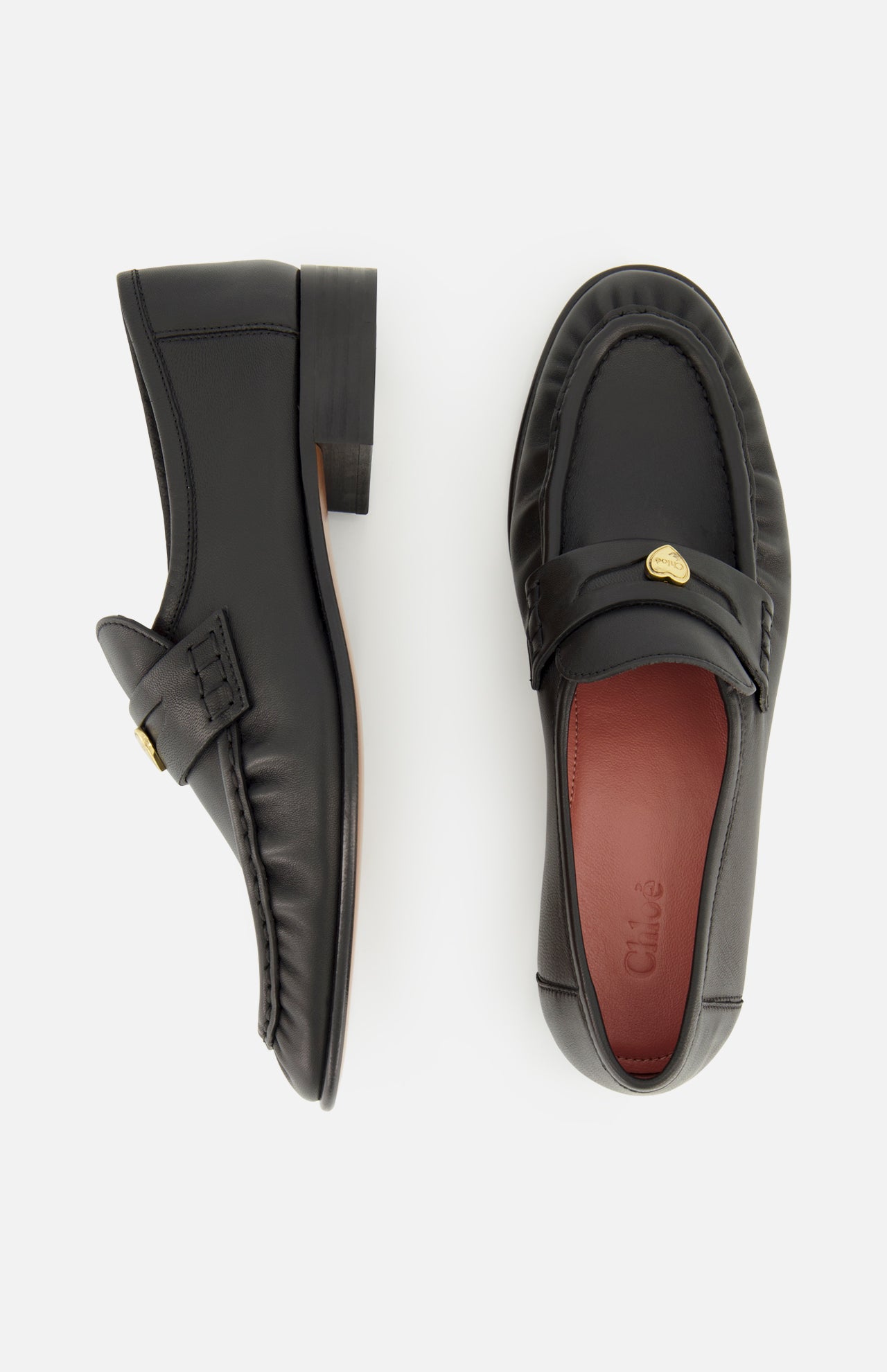 Two black Letti Gold Heart Loafers by CHLOÉ are displayed on a white background, with one shown from the side and the other from above. Each loafer features a gold-tone heart accent on the top, while the dark red insole bears the embossed CHLOÉ brand name. Combining classic style with luxurious detail, these loafers are an elegant choice for feminine flats.
