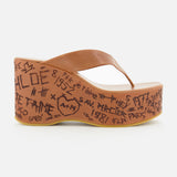 The Maxime Wedged Thong Sandals by CHLOÉ feature a brown wedge with a wood-effect platform decorated in black graffiti-style writing and symbols, complemented by a brown leather thong strap, shown against a white background.