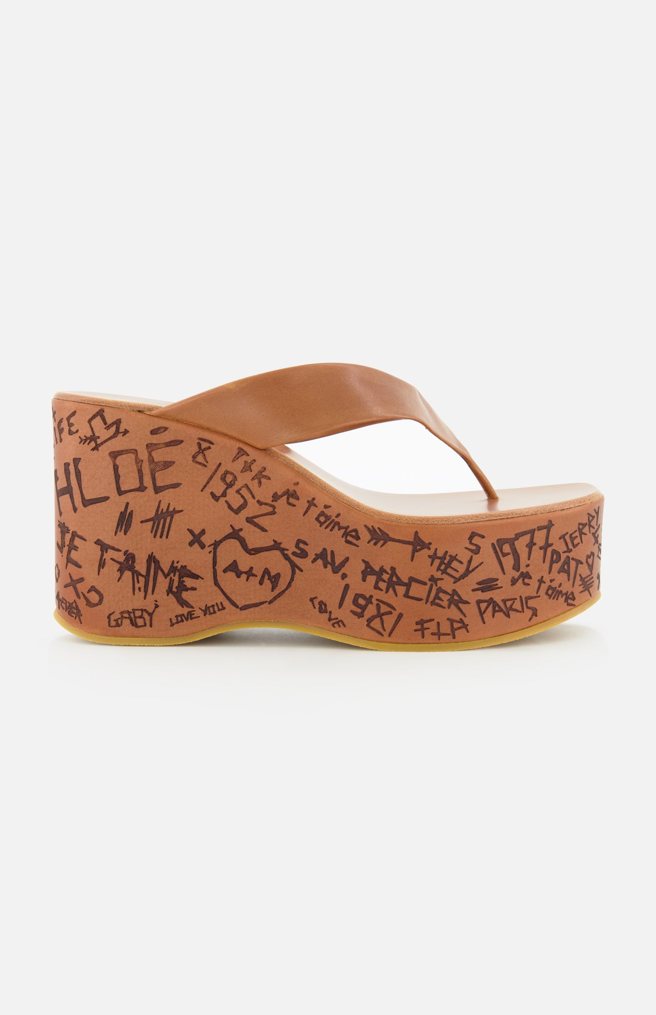 The Maxime Wedged Thong Sandals by CHLOÉ feature a brown wedge with a wood-effect platform decorated in black graffiti-style writing and symbols, complemented by a brown leather thong strap, shown against a white background.