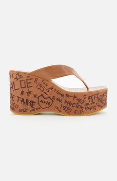 The Maxime Wedged Thong Sandals by CHLOÉ feature a brown wedge with a wood-effect platform decorated in black graffiti-style writing and symbols, complemented by a brown leather thong strap, shown against a white background.