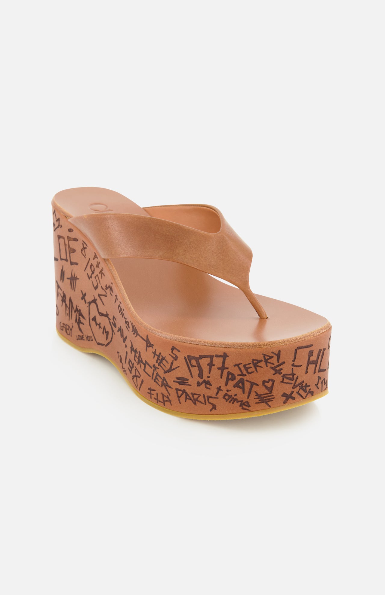 The Maxime Wedged Thong Sandals by CHLOÉ showcase brown leather straps and a thick, wood-effect platform wedge sole adorned with doodle-like black text and symbols along the side. Set against a plain white background.