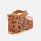The Maxime Wedged Thong Sandals by CHLOÉ are tan platform sandals featuring an open toe and decorated with graffiti-style black doodles and the word “CHLOE” along the outer side of their wood-effect platform and heel.