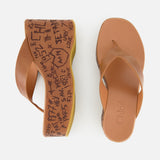 Two Maxime Wedged Thong Sandals by CHLOÉ, featuring a stylish brown leather thong design and thick, wood-effect platform soles adorned with black graffiti-style writing and symbols. The left sandal is displayed from above and the right from the side, both set against a plain white background. These wedges combine comfort with contemporary flair.