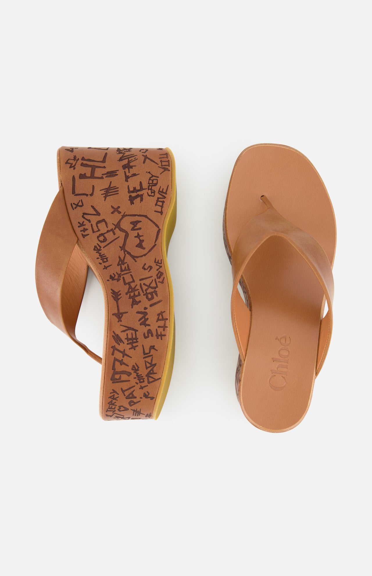 Two Maxime Wedged Thong Sandals by CHLOÉ, featuring a stylish brown leather thong design and thick, wood-effect platform soles adorned with black graffiti-style writing and symbols. The left sandal is displayed from above and the right from the side, both set against a plain white background. These wedges combine comfort with contemporary flair.