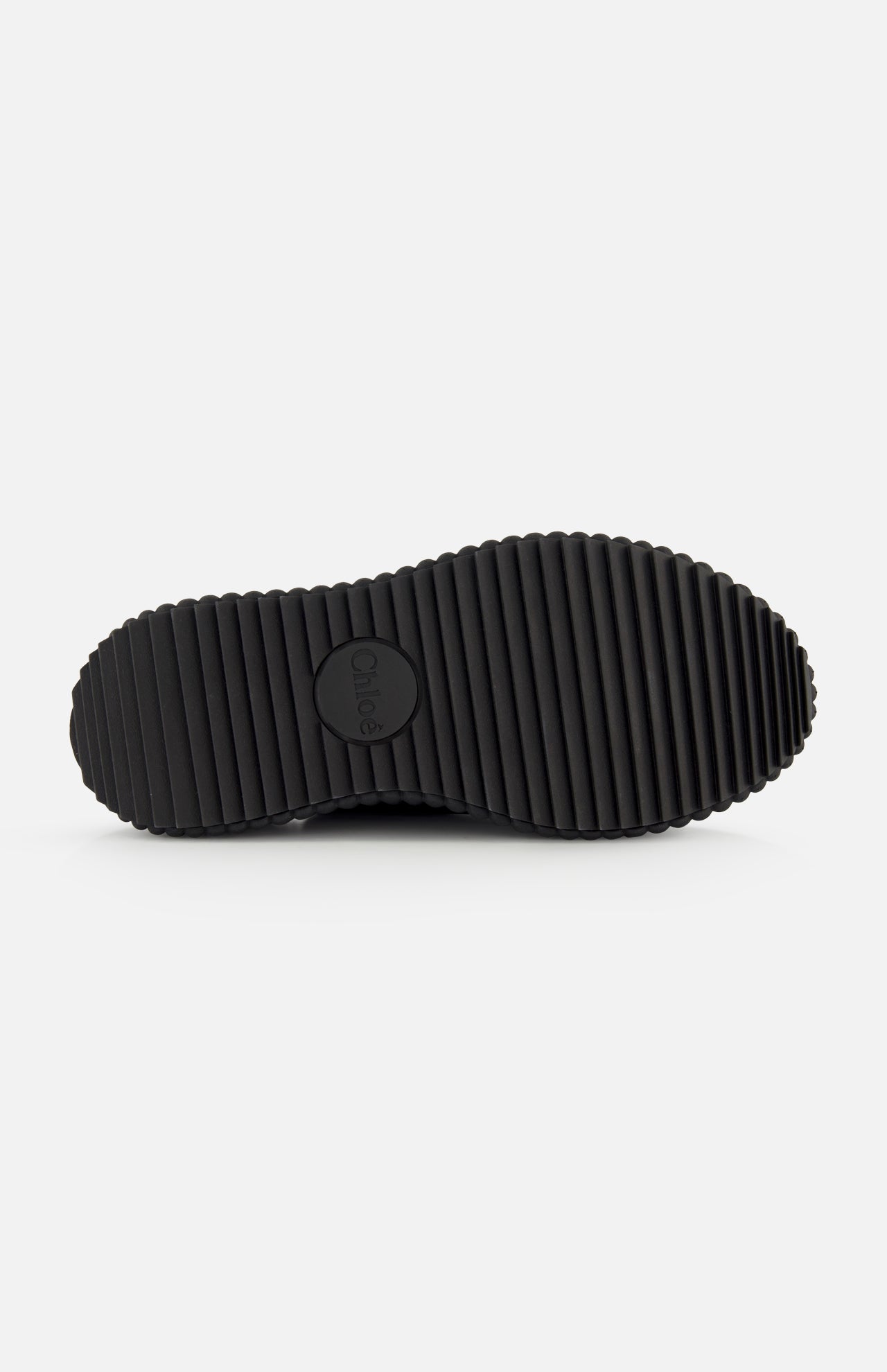 The image shows the underside of CHLOÉ’s Nama Buckle Wedge Sandals in black, featuring deep, evenly spaced horizontal grooves and a circular CHLOÉ logo at the center. The background is plain white.