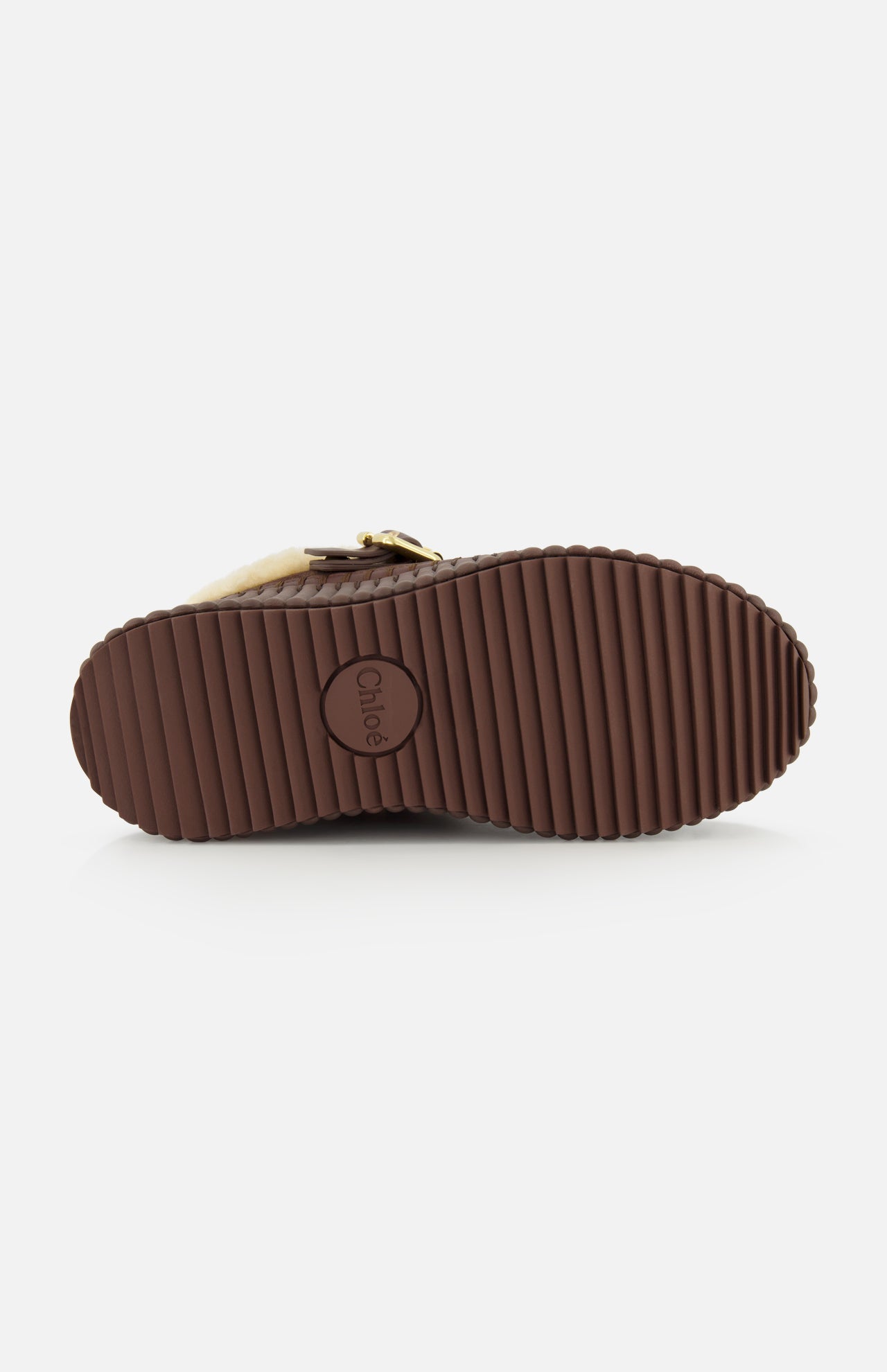 The image displays the ridged brown rubber sole of CHLOÉ’s Nama Suede Slippers, featuring the brand’s circular logo embossed at the center. A glimpse of the upper reveals soft light fur and a sleek strap, reflecting its plush shearling lining. This platform slip-on design effortlessly merges comfort and luxurious style.