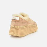 The Nama Shearling Lining Sneakers by CHLOÉ are beige slip-on shoes featuring a thick, ribbed recycled sole, visible stitching, and cream-colored shearling lining. Chloé branding appears on a white label at the heel.