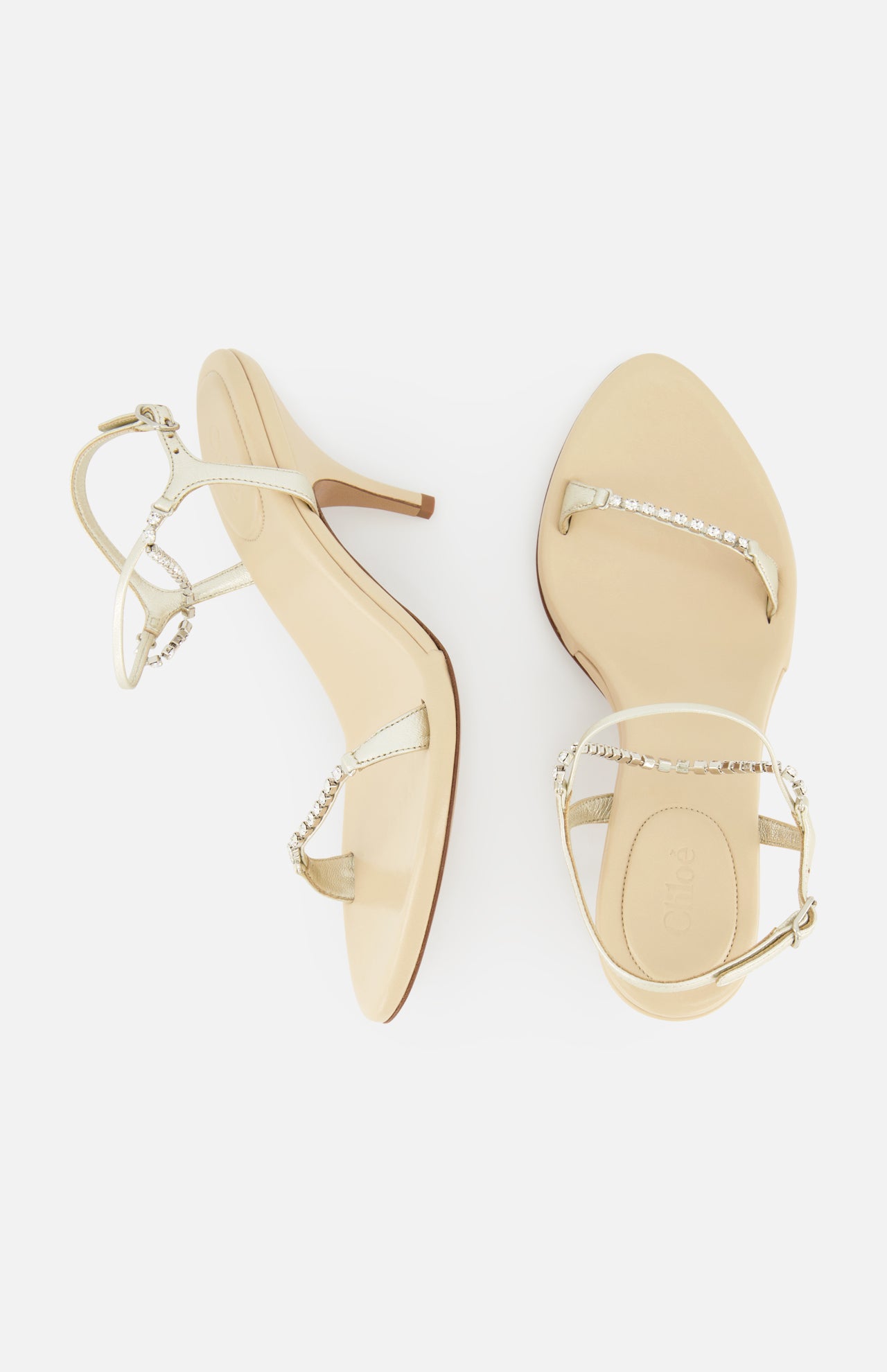A pair of Zia Crystal Chain Heels by CHLOÉ, featuring beige high heels with slender straps—one accentuated by a sparkling crystal chain—for an elegant interpretation of Italian designer footwear. Displayed on a white background, one shoe is shown from the top and the other from the side.