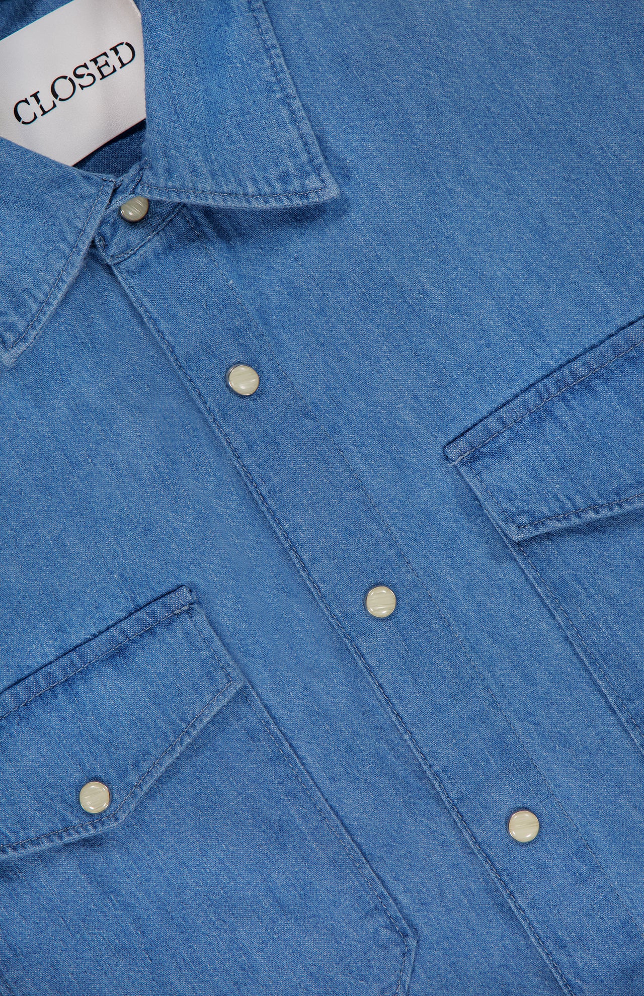 Close-up of the Army Long Sleeve Shirt by CLOSED, a regular fit blue denim shirt with chest patch pockets, silver snap buttons, and a collar. The inner collar features a white label with CLOSED in black text.