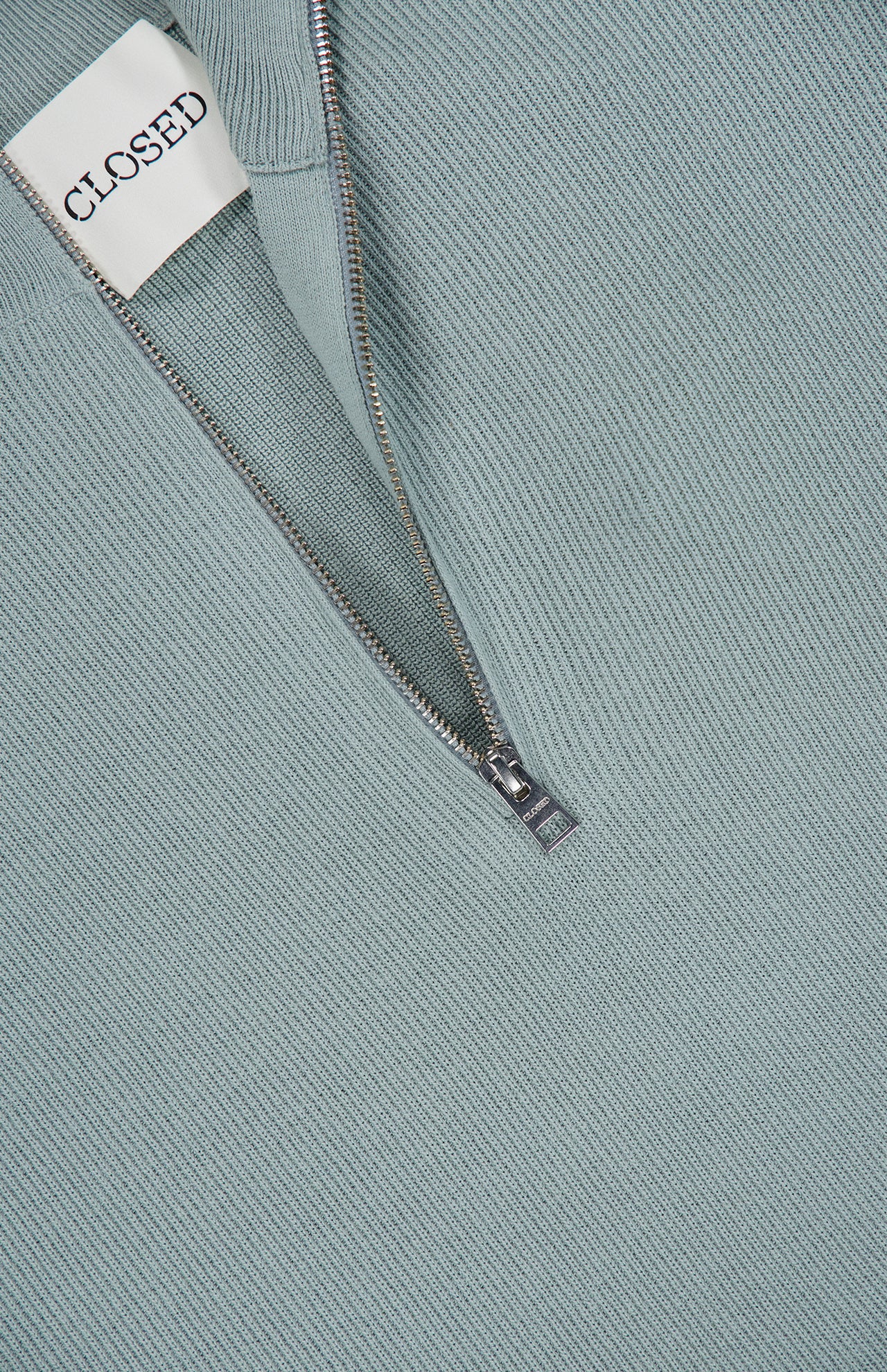 A close-up of the HALF ZIP JUMPER SWEATER by CLOSED in light teal cotton-blend fabric, featuring a partially unzipped silver zipper and a white label with the brand name CLOSED.