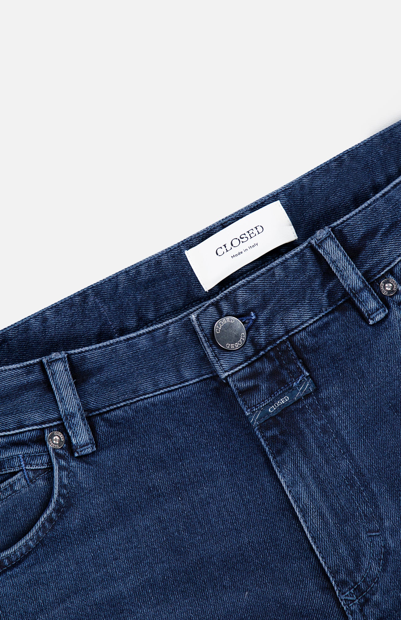 Close-up of the top part of CLOSED Unity Slim Jean in dark blue denim, featuring a metal button, belt loops, front pocket, and a white label with the CLOSED brand name stitched on the inside waistband. Expertly crafted from premium materials and made in Italy.
