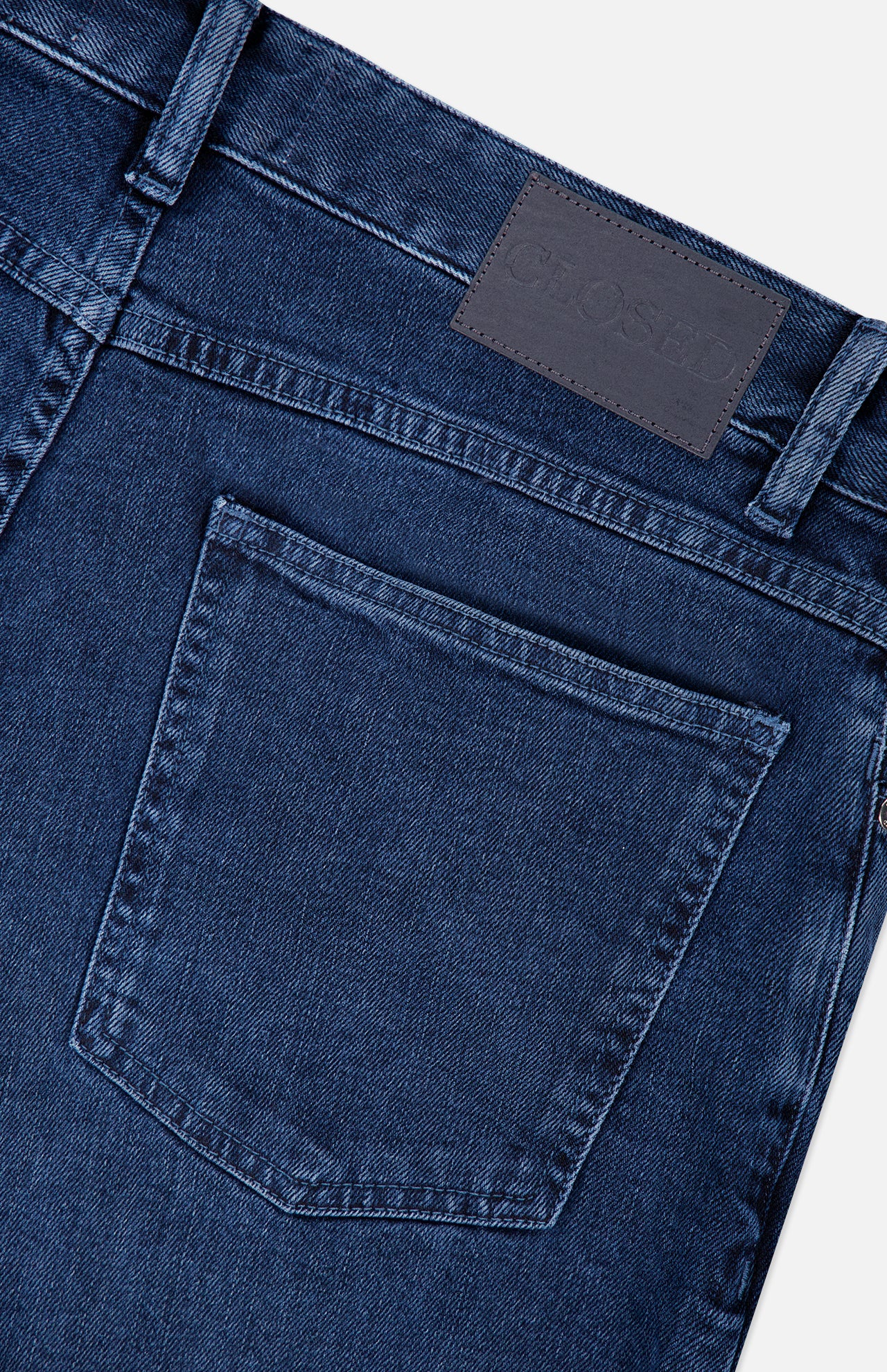 Close-up of the back pocket and waistband of the CLOSED Unity Slim Jean in dark blue denim, featuring a dark leather patch with embossed branding and visible stitching details.