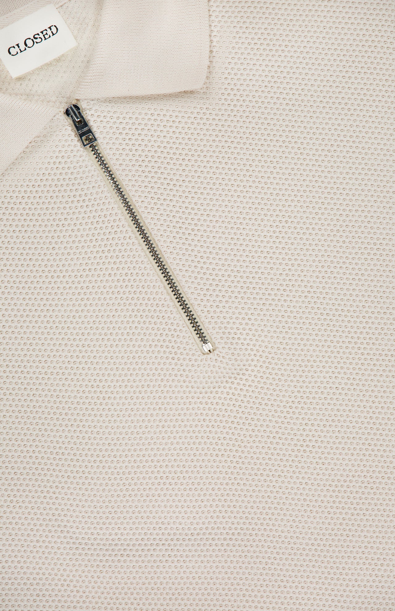 Close-up of the Zipped Neck Polo Shirt by CLOSED, featuring cream textured cotton, a partially open silver zip at the neck, and a white collar label with CLOSED in black uppercase letters.