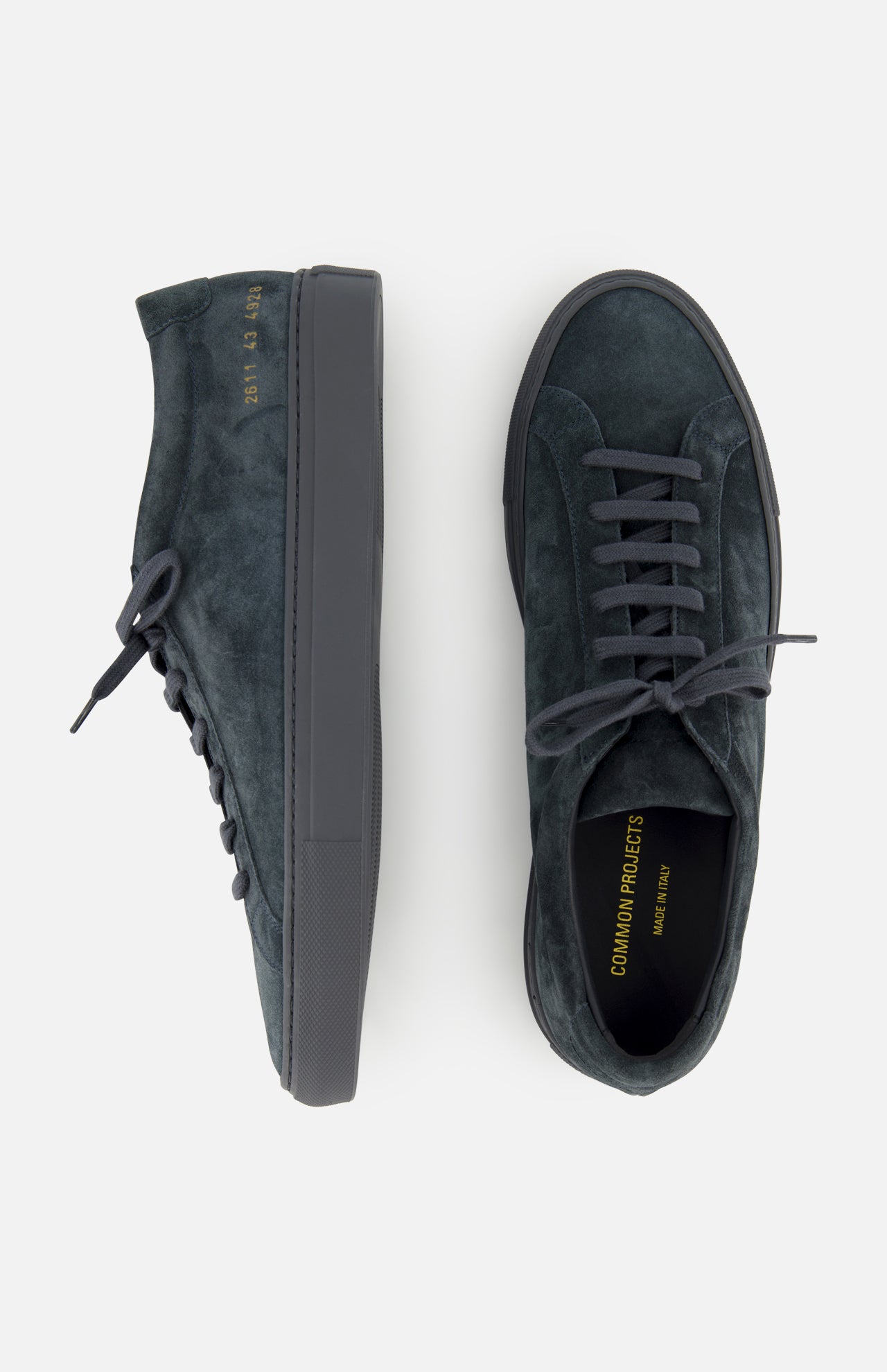 A pair of COMMON PROJECTS Achilles Suede Premium Sneakers in dark green suede features black soles and laces, shown from the side and top. The insoles are stamped with “COMMON PROJECTS” and “MADE IN ITALY” in gold lettering, emphasizing their luxurious low-top design.