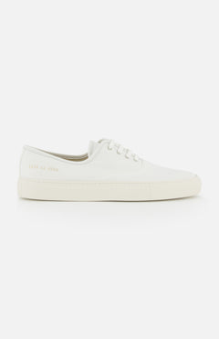The COMMON PROJECTS Four Hole Slip On In Canvas Sneaker features a minimalist white design with white laces, an off-white rubber sole, and a small gold serial number on the heel, set against a simple light background.
