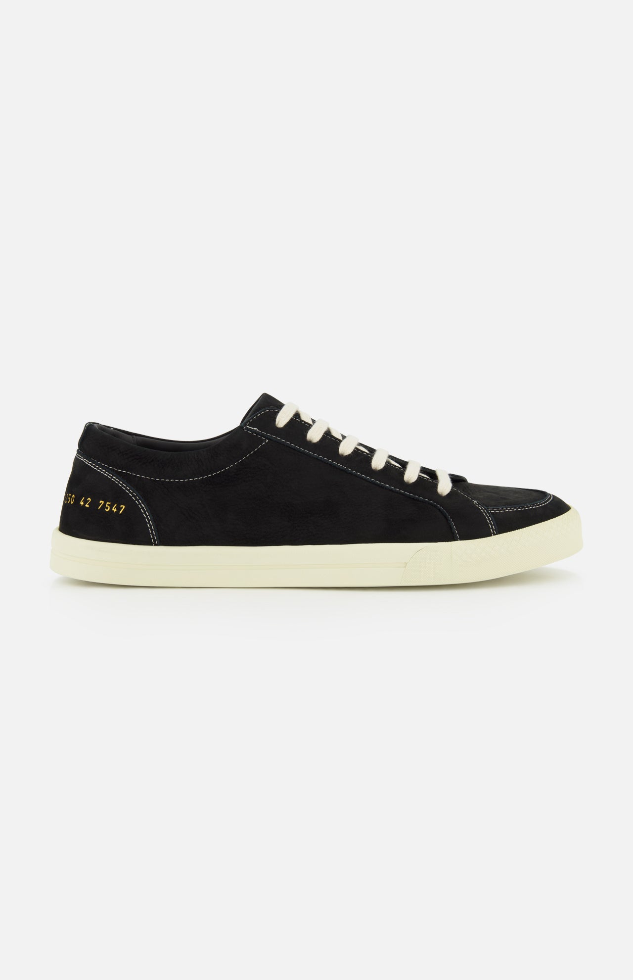 The COMMON PROJECTS Premium Skate In Nubuck Sneaker is a black nubuck low-top with white laces and a crisp white sole, displayed from the side. Signature gold numbers stamped near the heel lend an exclusive touch to this luxury designer shoe, while visible white stitching highlights its refined construction.