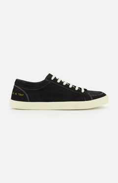 The COMMON PROJECTS Premium Skate In Nubuck Sneaker is a black nubuck low-top with white laces and a crisp white sole, displayed from the side. Signature gold numbers stamped near the heel lend an exclusive touch to this luxury designer shoe, while visible white stitching highlights its refined construction.