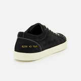 A rear view of the COMMON PROJECTS Premium Skate In Nubuck Sneaker showcases its black nubuck construction with white laces and a matching white sole. Gold numbers 5250 42 7547 are stamped near the heel, emphasizing its premium low-top design. The sneaker is set against a simple light gray background.