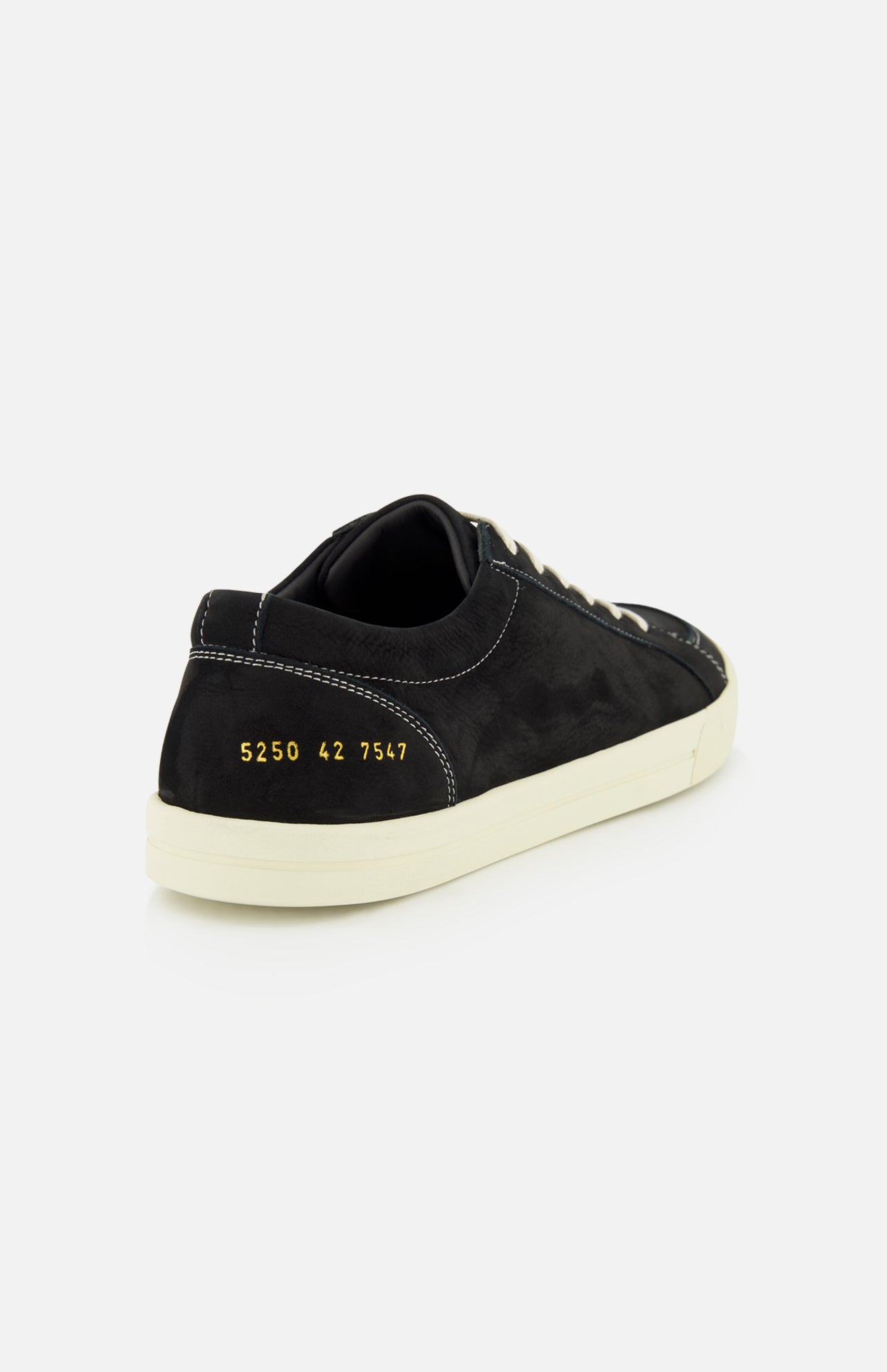 A rear view of the COMMON PROJECTS Premium Skate In Nubuck Sneaker showcases its black nubuck construction with white laces and a matching white sole. Gold numbers 5250 42 7547 are stamped near the heel, emphasizing its premium low-top design. The sneaker is set against a simple light gray background.