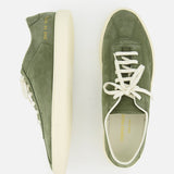 The Leather Tennis Sneaker by COMMON PROJECTS is an olive green suede luxury street shoe with white laces, cream rubber soles, and gold embossed branding on the side and insole. Shown from side and top view on a white background.