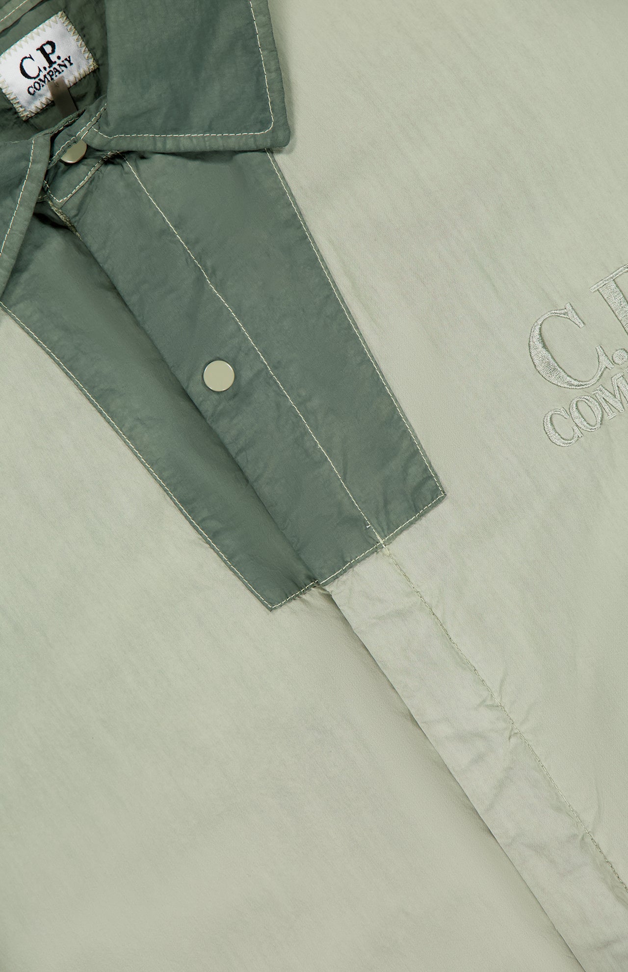Close-up of the C.P. COMPANY 50 Fili Two-tone Overshirt in moss green, featuring snap buttons, visible white stitching, and an embroidered logo for a refined utilitarian look.