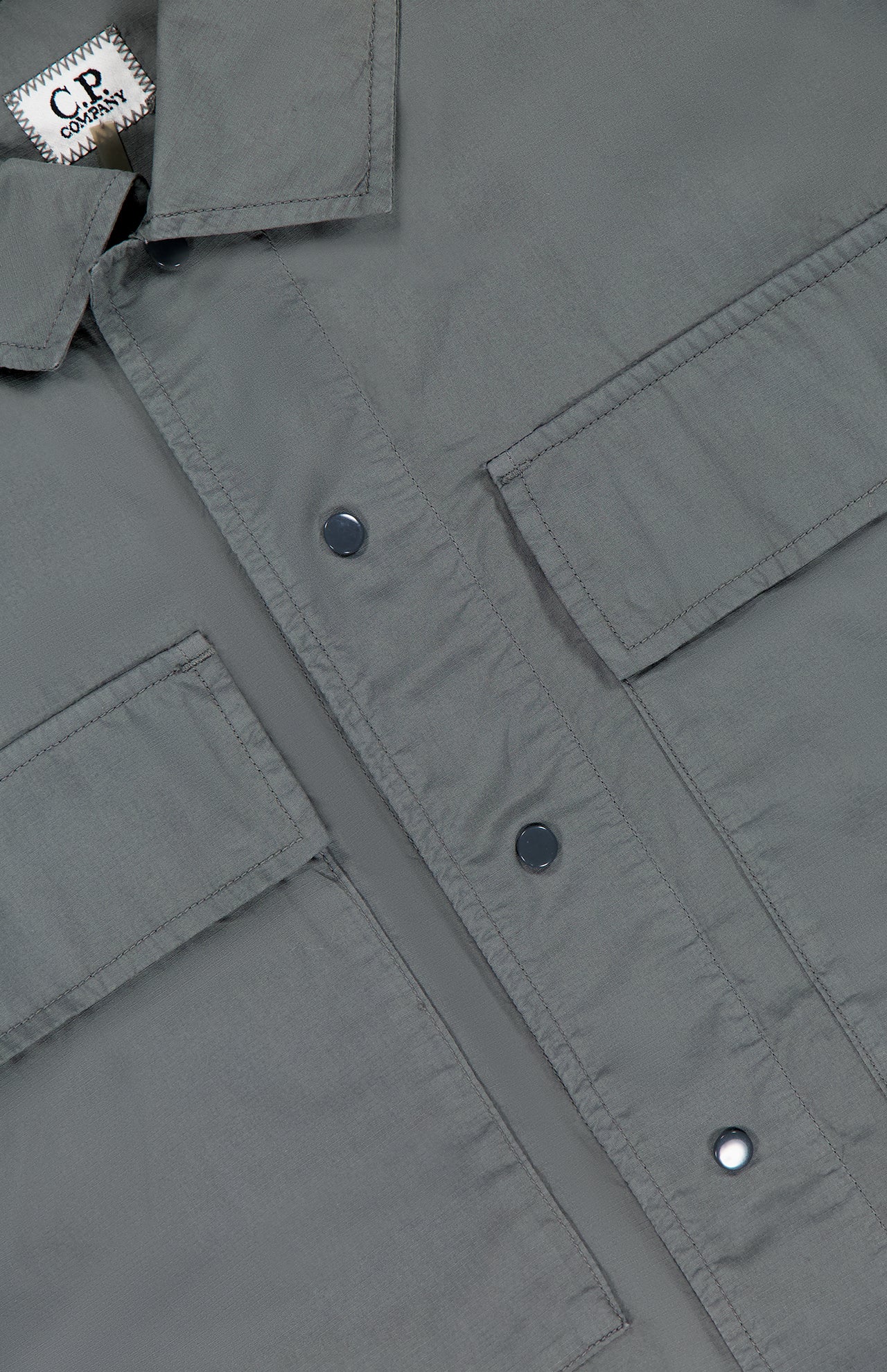 Close-up of the Cotton Rip-Stop Overshirt by C.P. COMPANY in grey, showcasing two large front pockets, metal snap buttons, and smooth 100% cotton fabric with a visible white label at the top left.