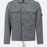 The C.P. COMPANY Cotton Rip-Stop Overshirt is a grey, 100% cotton jacket with front snap buttons, two chest pockets, a small left sleeve pocket, and a drawstring hem. Shown on a white background.