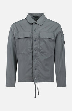 The C.P. COMPANY Cotton Rip-Stop Overshirt is a grey, 100% cotton jacket with front snap buttons, two chest pockets, a small left sleeve pocket, and a drawstring hem. Shown on a white background.