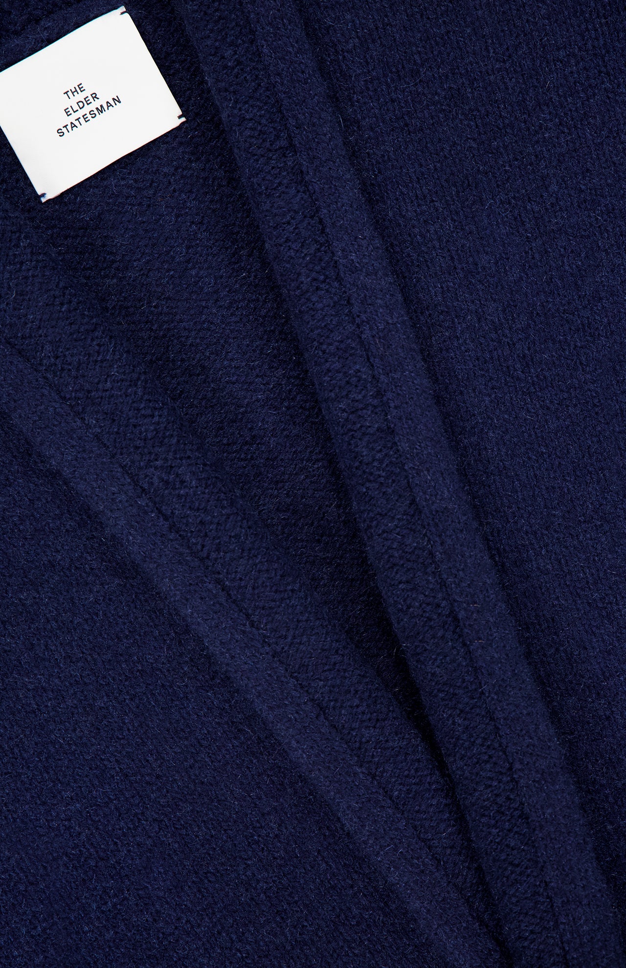 Close-up of a dark blue textured fabric with visible seams and a white label that reads ELDER STATESMAN, evoking the luxurious Italy Cashmere Smoking Cardigan with its refined shawl collar.