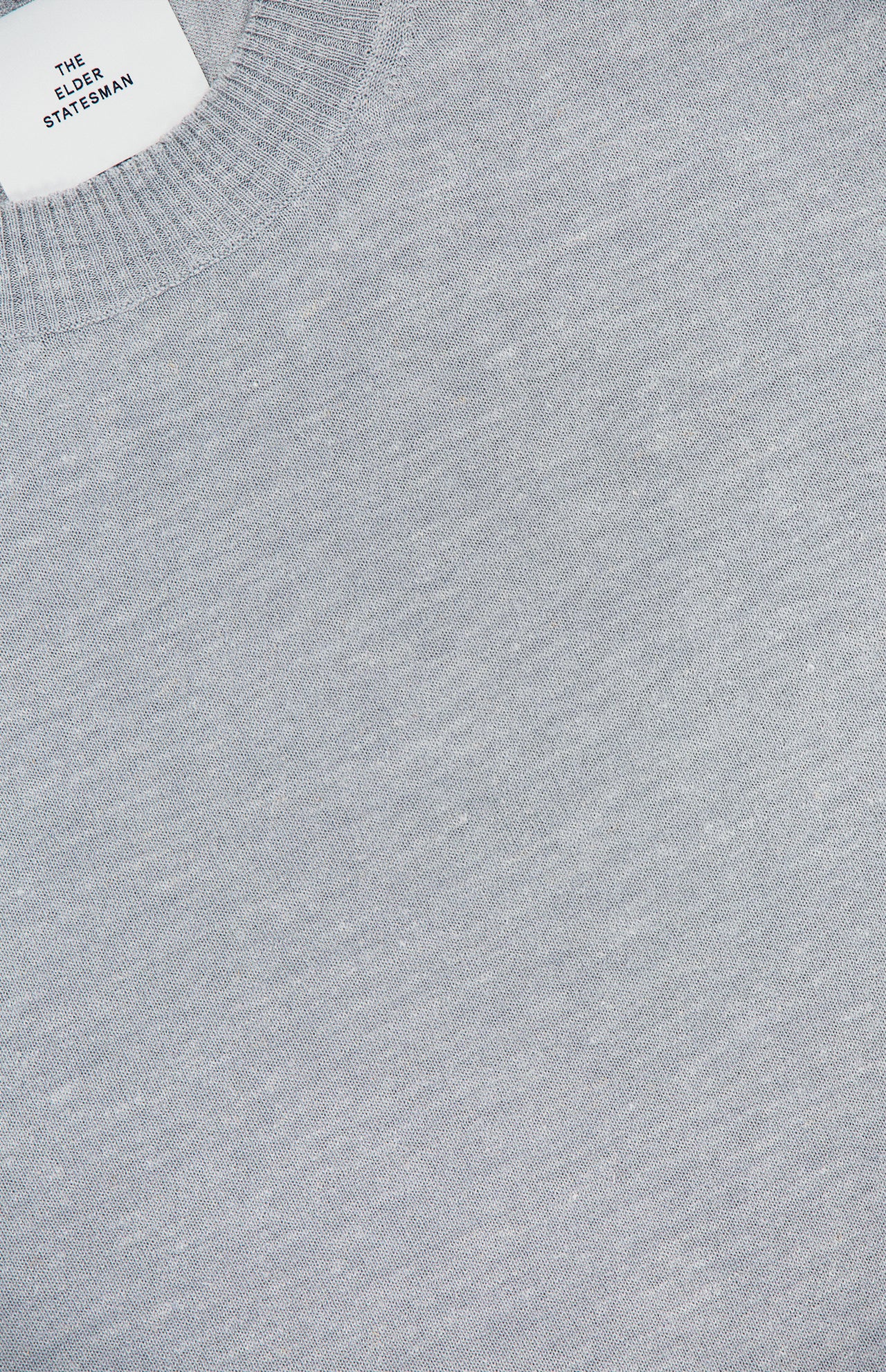 Close-up of the Libeccio Wren Crewneck Sweater in light gray, featuring a ribbed collar and expertly crafted from a luxurious blend of wool, silk, and linen. A white label with black text reading “ELDER STATESMAN” is partially visible at the neckline, highlighting the refined craftsmanship associated with this premium knitwear brand.