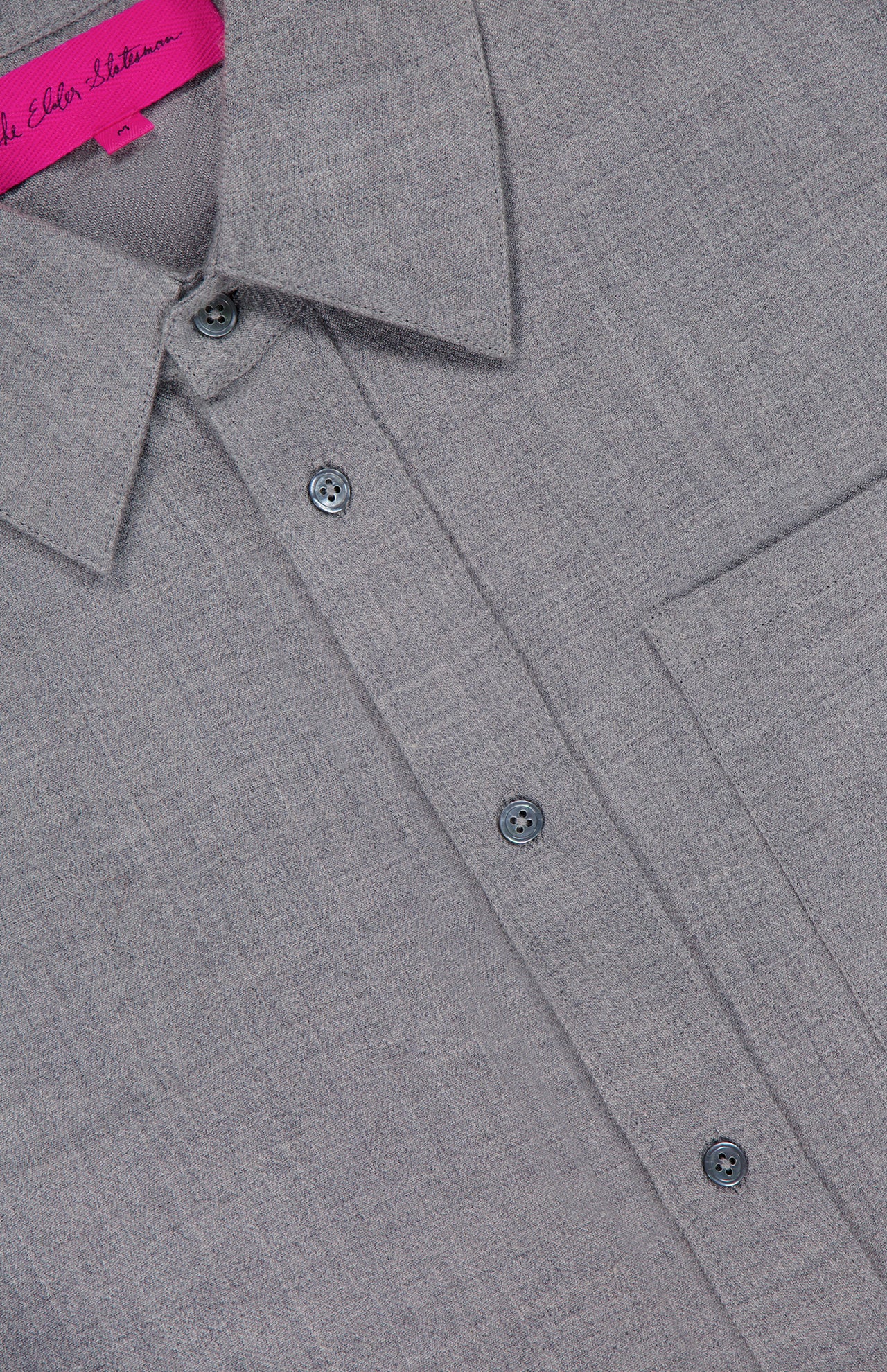 Close-up of the Pashmina Big Button Up Shirt by ELDER STATESMAN in light grey, featuring a classic collar, chest pocket, and four visible buttons. A partially visible pink label with text appears at the neckline. Designed as an ideal piece of office wear, the shirt's fabric boasts a soft, textured look reminiscent of luxurious wool cashmere.