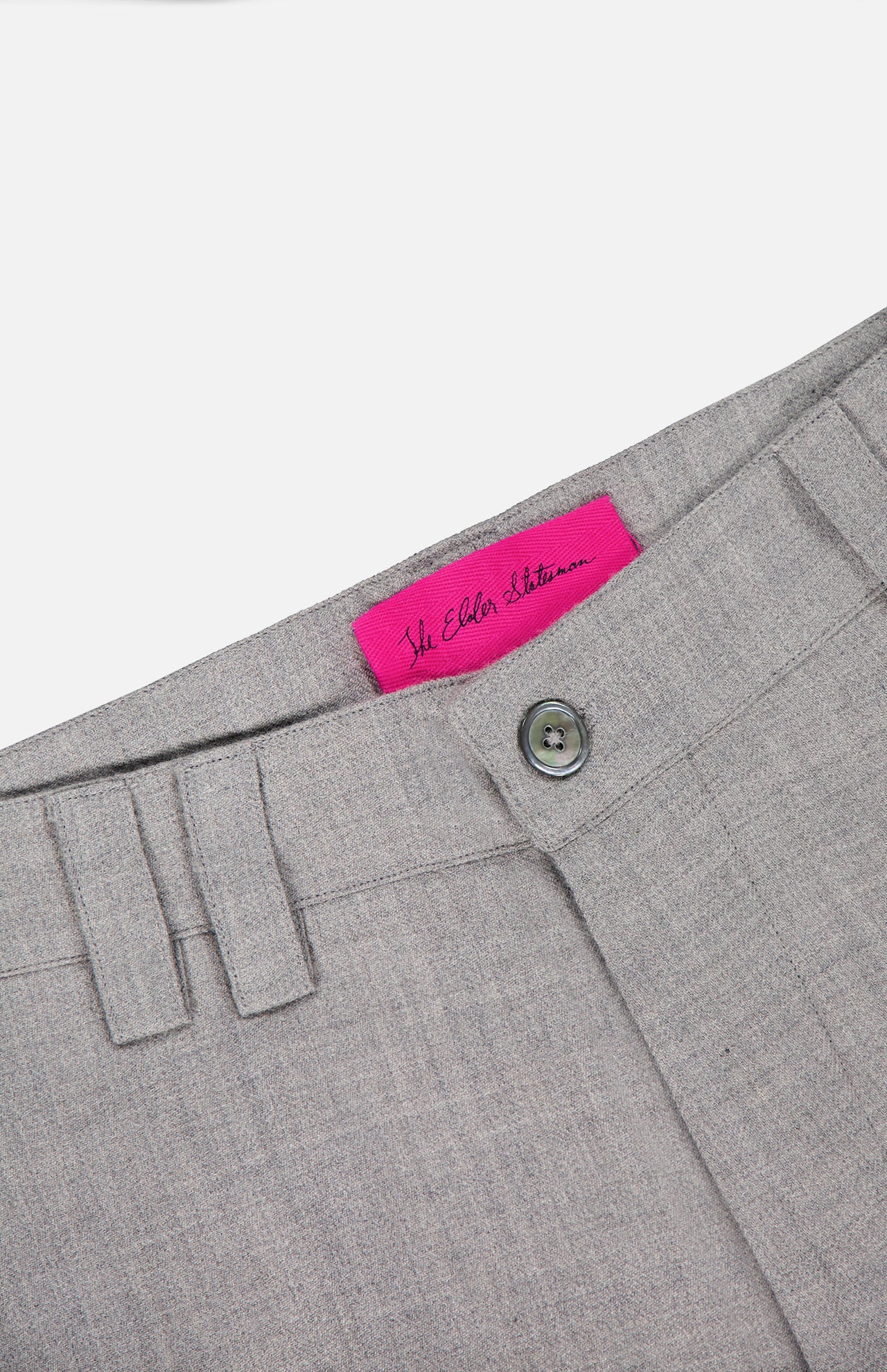 Close-up of the waistband of the ELDER STATESMAN Pashmina Norm Pant in grey, highlighting the button and belt loops, with a bright pink label featuring cursive writing on the inside.