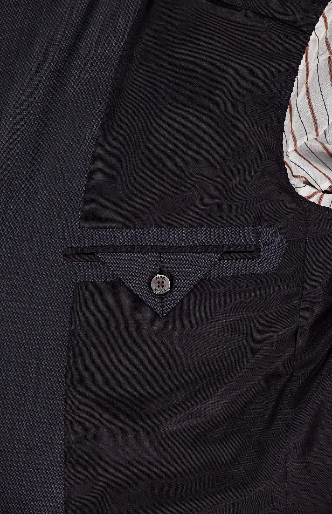 Close-up of the ZEGNA 15MilMil15 Wool Suit's dark grey interior lining, made in Italy, showing a neatly stitched pocket with a triangular flap and button. A striped fabric lining is visible on the right.