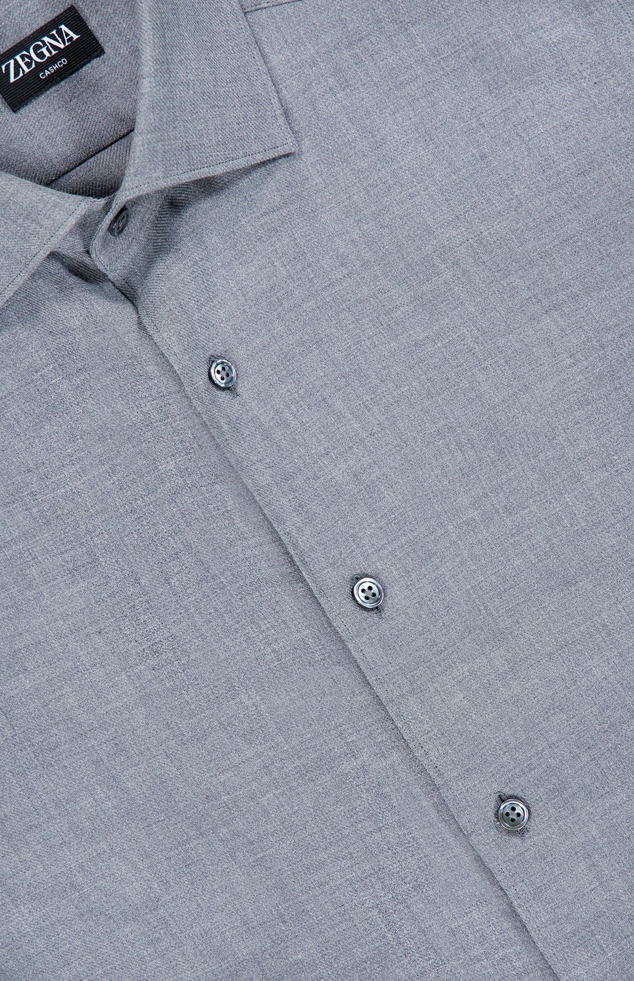 Close-up of the ZEGNA Cashco Dress Shirt in light grey textured fabric, showcasing a pointed collar and three visible buttons. The black label features “ZEGNA” and “Como” in white text, emphasizing this refined cashmere-cotton shirt crafted in Italy.