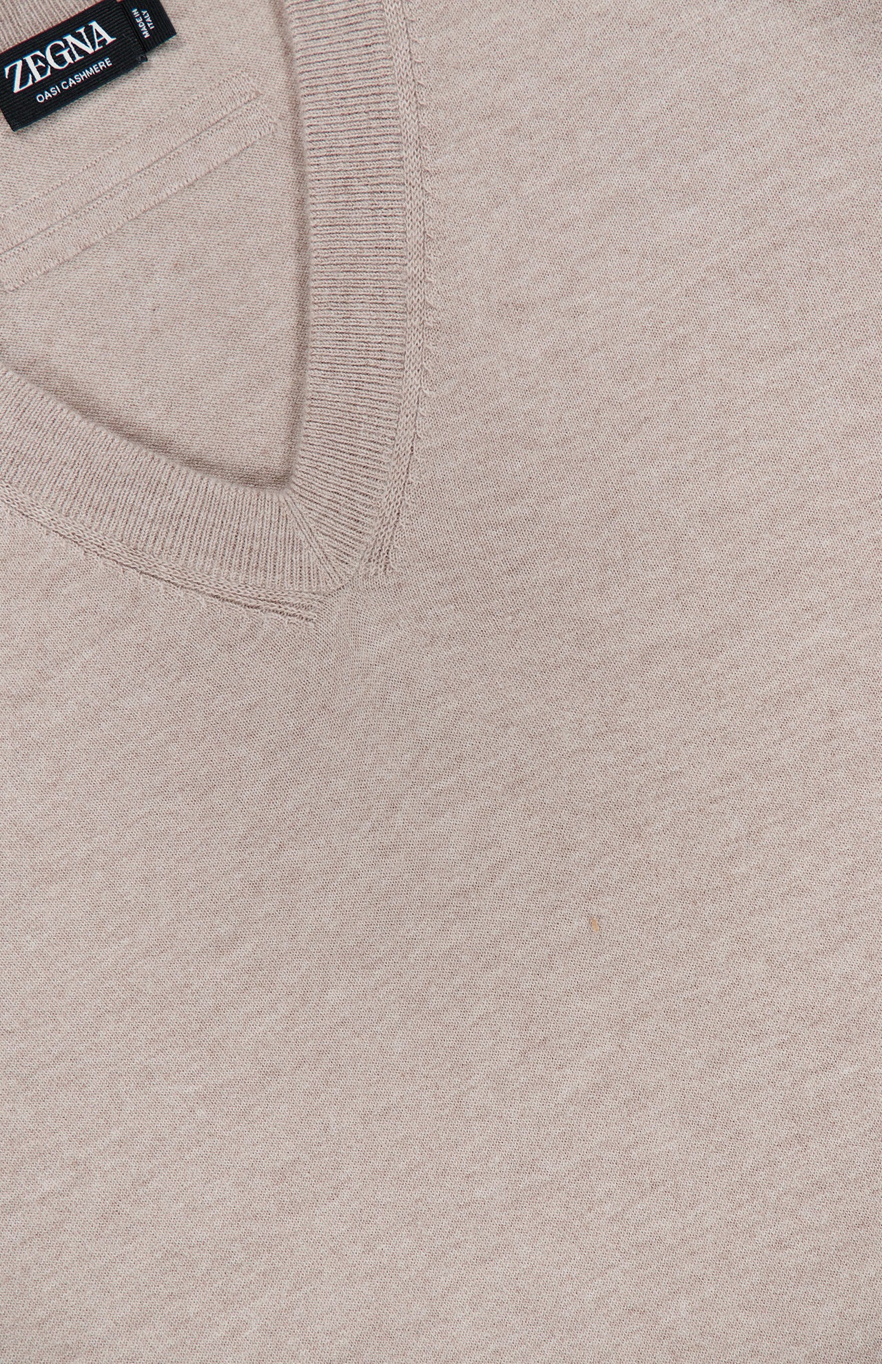 Close-up of the Oasi Cashmere V-Neck Sweater by ZEGNA in beige, highlighting its fine knit texture and featuring a black ZEGNA label at the upper left; this luxurious sweater is expertly crafted in Italy from premium cashmere.