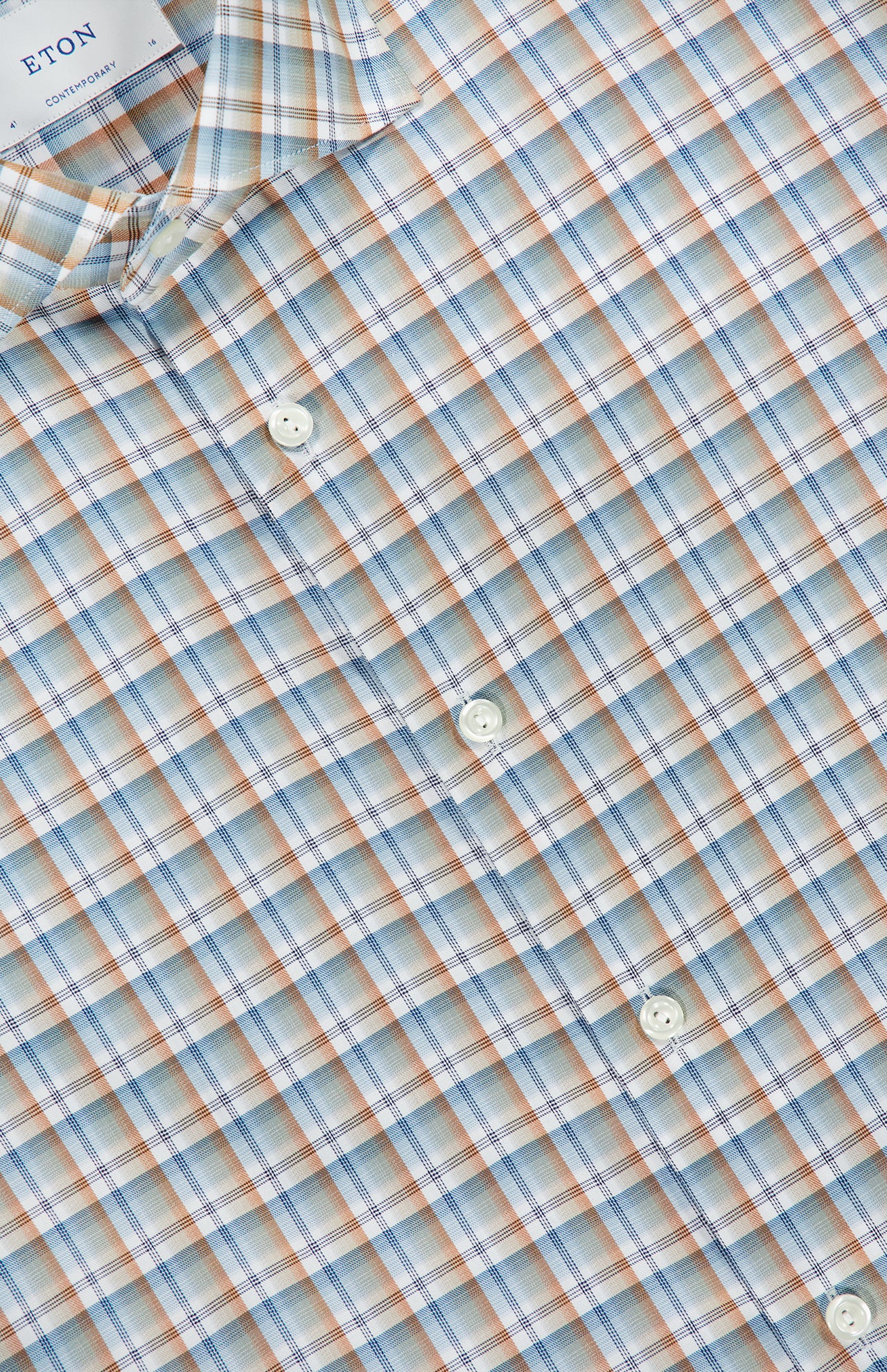 Close-up of the ETON Check Twill Contemporary Dress Shirt in light blue, featuring a subtle orange and white plaid pattern. The collar displays an ETON fabric label. Made from organic cotton, this dress shirt combines style and sustainability with four visible white buttons on the front.